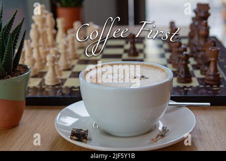 Eine Tasse schaumigen Kaffee und schwarz-weiße Schachfiguren Reihen sich auf einem Brett im Hintergrund an. Inschrift: KAFFEE ZUERST oben im Bild. Stockfoto