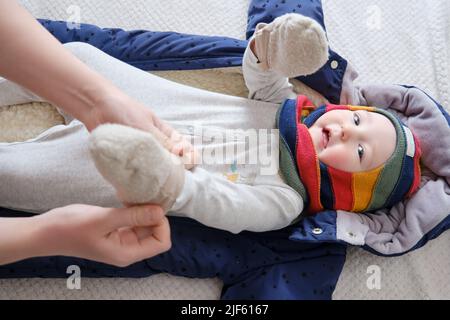 Eine Mutter legt warme Fäustlinge für einen kleinen Jungen an. Mama kleidet ein glückliches Kind in Winterkleidung auf dem Bett. Stockfoto