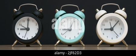 Drei kleine Wecker schwarz blau und weiß zeigen neun Uhr, stehen auf einem Tisch auf schwarzem Hintergrund Stockfoto