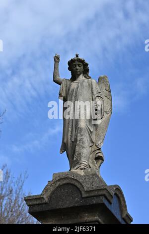 Abgenutzte Steinskulptur eines Engels, der beim Blick nach unten in den Himmel zeigt, mit blauem Himmel und hellen Wolken im Hintergrund Stockfoto