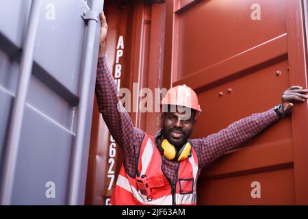 Glücklicher afroamerikanischer Ingenieur lächelt, als er im Containerlager steht, Arbeiter bekommen Sozialversicherung und Versicherung, Labor Day und Erfolg Stockfoto