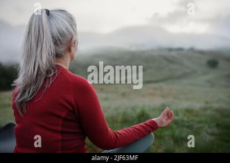 Rückansicht einer älteren Frau, die am frühen Morgen in der Natur Atemübungen mit Nebel und Bergen im Hintergrund macht. Stockfoto