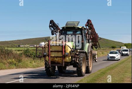 Cornwall, England, Großbritannien. 2022. Grüner Traktor mit montiertem Chemikalientank und Spritzgerät auf dem Heck, der entlang einer Landstraße in der Nähe von St fährt Stockfoto