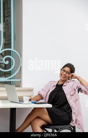 Lächelnde lateinische junge Frau mit Laptop, während sie im Straßencafé sitzt - Stockfoto Stockfoto