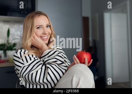 Nahaufnahme eines charmanten jungen lächelnden Blondinen, die sich wohlfühlt wie zu Hause und einen saftigen roten Apfel in den Händen hält. Stockfoto