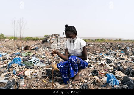 Afrikanisches Mädchen, das inmitten von Abfallhaufen sitzt und für die Suche nach recycelbaren Materialien wie Metall, Glas und Kunststoff verantwortlich ist; informelle Abfallrezyk Stockfoto