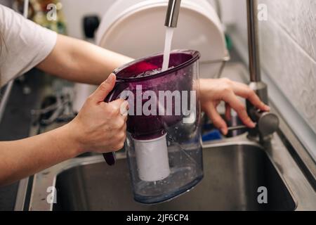 Frau goss Wasser aus dem Wasserhahn in den Wasserfilterkrug in der Küche. Gesunder Lebensstil. Frau, die Wasser füllte. Stockfoto