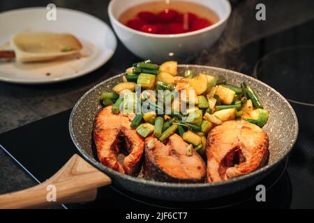 Nahaufnahme einer unkenntlichen Frau, die das Mittagessen in der Küche zubereitet. Gegrilltes Forellensteak. Braten unter Rühren. Die Zucchini und die grünen Bohnen werden in der Pfanne gebraten. Strom Stockfoto