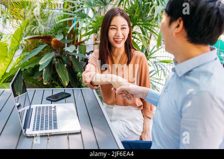 Erfolgreicher junger asiatischer Geschäftsmann und Geschäftsfrau, die sich die Hände schüttelten, um den Deal zu bestätigen und einen Laptop zu benutzen, während sie sich im modernen Café treffen Stockfoto