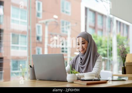 Junge asiatische muslimische Geschäftsfrau in smarter Casual-Kleidung diskutieren Geschäft und lächeln, während sie im kreativen Coworking sitzt. Stockfoto