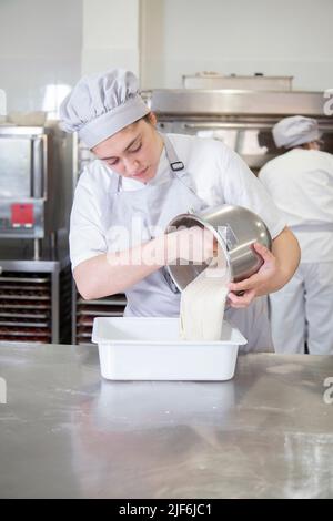 Seitenansicht einer Bäckerin in Uniform, die rohen Teig aus der Schüssel in den Behälter legt, während sie in einer professionellen leichten Bäckerei arbeitet Stockfoto
