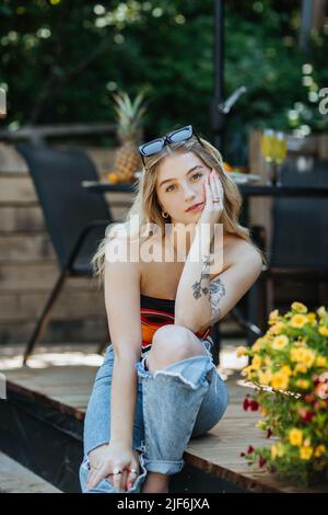 Attraktive junge Frau in legerer Kleidung mit Tätowierungen am Arm und Blick auf die Kamera, während sie am Sommertag auf der Terrasse des Gebäudes sitzt Stockfoto