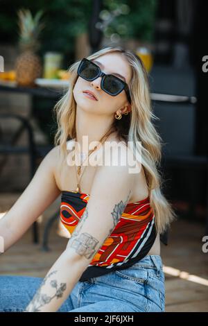 Attraktive junge Frau in legerer Kleidung mit Tätowierungen am Arm und Blick auf die Kamera, während sie am Sommertag auf der Terrasse des Gebäudes sitzt Stockfoto