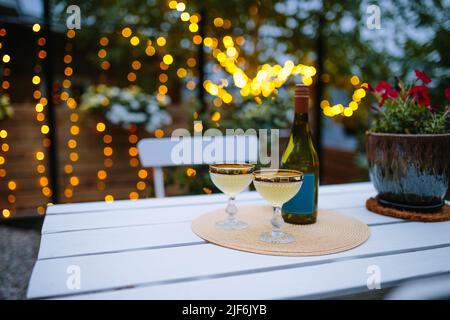 Gläser mit Champagner auf weißem Holztisch mit Flasche auf der Terrasse des Gebäudes mit Bäumen vor verschwommenem Hintergrund serviert Stockfoto