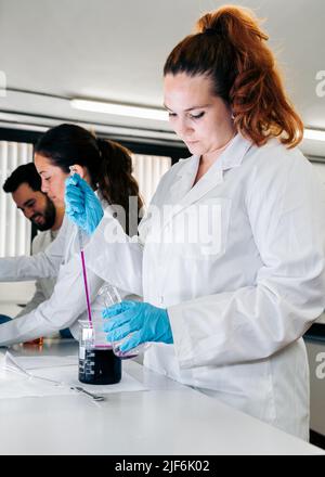 Konzentrierte Wissenschaftlerin in medizinischer Uniform, die in der Nähe von Kollegen steht und dabei bunte chemische Flüssigkeit aus der Pipette in den Glaskolben gießt Stockfoto