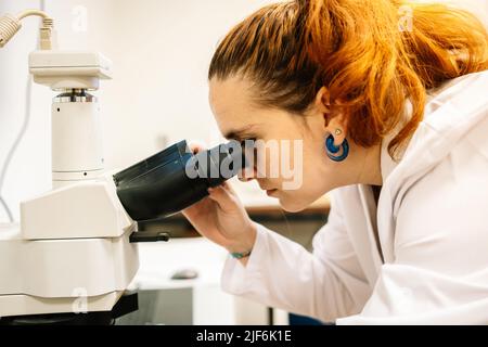 Seitenansicht einer konzentrierten Wissenschaftlerin in weißer medizinischer Uniform, die durch das moderne Mikroskop schaut, während sie Proben im Lichtlabor untersucht Stockfoto
