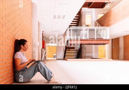 Ganzkörper-Seitenansicht einer entzückten Studentin in Freizeitkleidung, die auf Netbook im Internet surft, während sie auf dem Boden in der Nähe einer Ziegelwand sitzt Stockfoto