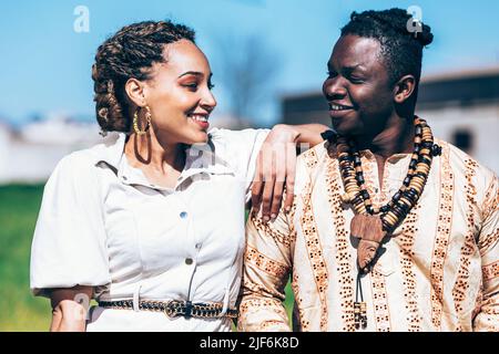 Mixed Race Frau in lässigem weißen Kleid auf der Schulter des glücklichen schwarzen Mannes in traditionellen afrikanischen Hemd mit Halsketten lehnt, während in grünen Gras stehen Stockfoto
