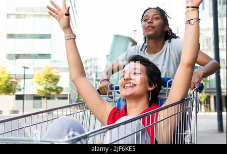 Afroamerikanische Frau, die mit einer fröhlichen jungen Freundin einen Einkaufswagen aus Metall schiebt, während sie sich auf der Straße der Stadt amüsieren Stockfoto