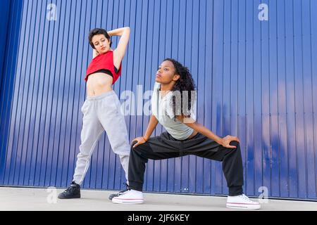 Ein ganzer Körper von sportlichen, vielfältigen Tänzerinnen in aktiven Trainingseinheiten, die sich vor dem Training auf der Straße in der Nähe der blauen Mauer in der Stadt zusammenstrecken Stockfoto