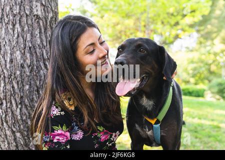 Fröhliche weibliche Besitzerin mit langen Haaren lächelnd und liebkochend loyaler Labrador Retriever Hund mit schwarzem Fell, während er sich am Sommertag in der Nähe des Baumstammes ausruhte Stockfoto