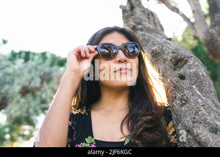 Frau in Sonnenbrille, die in hinterleuchteter Beleuchtung auf dem Baumstamm im Park zurückblickt Stockfoto