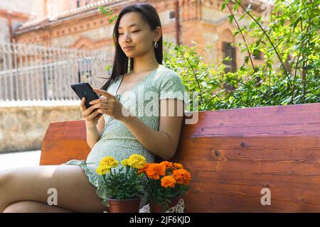 Charmante asiatische weibliche SMS-Nachricht auf Handy, während auf der Bank mit bunten Ringelblumen Topfblumen im städtischen Garten sitzen Stockfoto