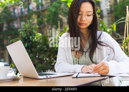 Attraktive asiatische Freiberuflerin, die Notizen im Notizblock macht, während sie an einem Projekt am Tisch mit Laptop auf der Terrasse der Cafeteria in der Stadt arbeitet Stockfoto