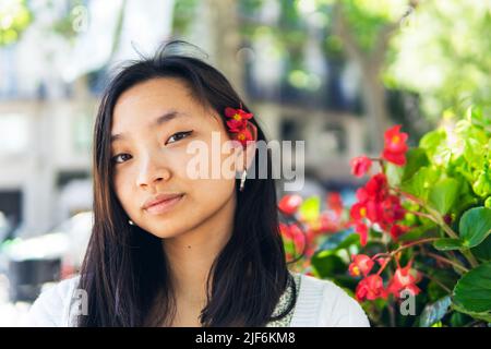 Attraktive junge asiatische Frau mit roten Blüten hinter dem Ohr, die im Sommer vor verschwommenem Hintergrund auf die Kamera auf der sonnigen Straße blickt Stockfoto