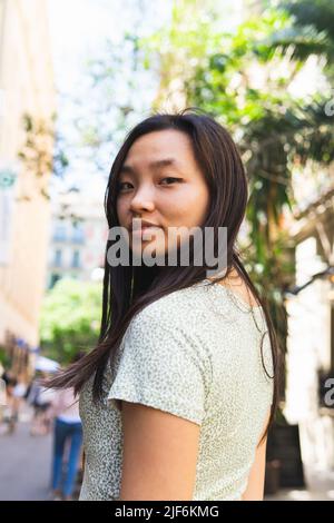 Seitenansicht einer jungen asiatischen Frau, die im Sommer auf der Straße der Stadt auf die Kamera schaut Stockfoto