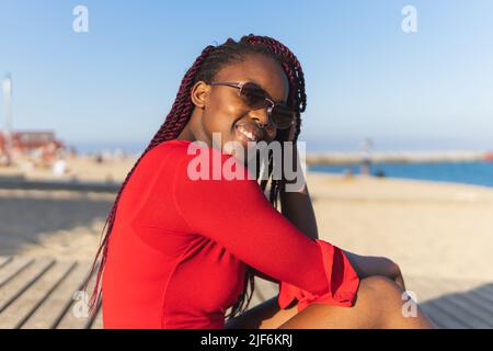 Porträt einer afroamerikanischen jungen Frau in trendigem roten Kleid und Zöpfen, die beim Sitzen am Strand auf die Kamera schauen Stockfoto