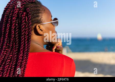 Rückansicht der afroamerikanischen jungen Frau in trendigem roten Kleid und Zöpfen, die beim Sitzen am Strand auf das Meer blicken Stockfoto