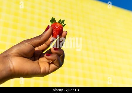 Hand einer anonymen afroamerikanischen Frau, die an einem sonnigen Tag Erdbeere vor gelbem Hintergrund hält Stockfoto