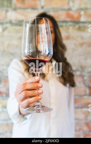 Weicher Fokus einer nicht erkennbaren Sommelierin in weißem Hemd, der im Restaurant einen Glaskelch aus Rotwein an der Backsteinmauer zeigt Stockfoto