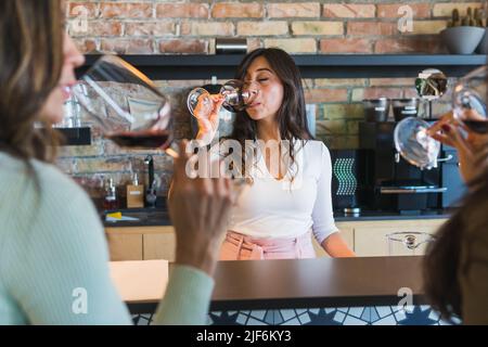 Die ethnische Barkeeperin in trendigen Kleidern schlürft Rotwein aus Glas, während sie mit Kollegen in der Bar Alkohol verkostet Stockfoto
