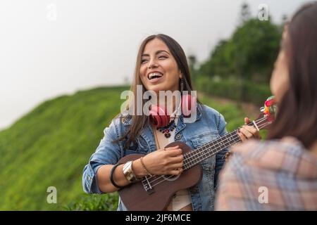 Fröhliche junge ethnische Frau mit langen dunklen Haaren in legerer Kleidung und Kopfhörern am Hals lächelnd, während sie Ukulele spielt und in der Natur mit Getreide singt Stockfoto