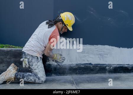 Seitenansicht eines professionellen Malers in gelbem Hardhut, der während der Verputzung der Wand auf dem Bürgersteig auf der Stadtstraße sitzt Stockfoto