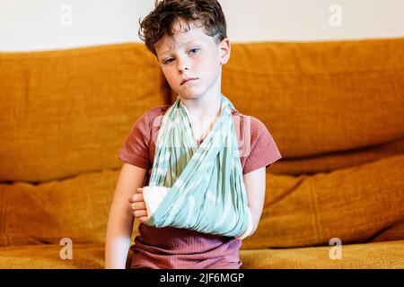 Kleiner Junge im T-Shirt streichelt gebrochenen Arm in einem Tragetuch und schaut auf die Kamera, während er auf dem Sofa sitzt und unter starken Schmerzen im Wohnzimmer zu Hause leidet Stockfoto