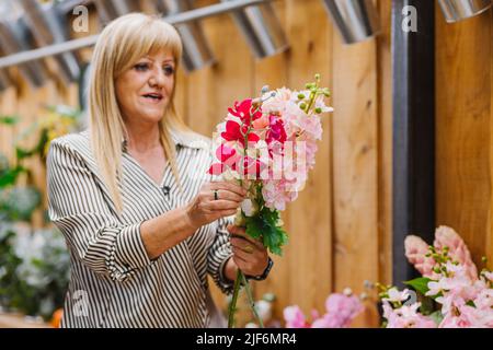 Ältere Frau in eleganter Freizeitkleidung mit langen blonden Haaren, die während der Arbeit im Geschäft frische Blumen für den Blumenstrauß auswählen Stockfoto