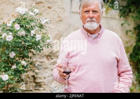 Älterer bärtiger Mann mit grauem Haar, der während der Hochzeitsfeier in der Nähe des Gebäudes Rotwein trinkt, gegen Menschen vor verschwommenem Hintergrund Stockfoto