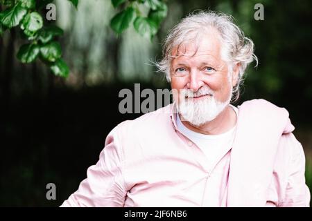 Älterer bärtiger Mann mit grauem Haar in rosa Hemd, der während der Festveranstaltung auf dem Land auf unscharfem Hintergrund steht und wegschaut Stockfoto