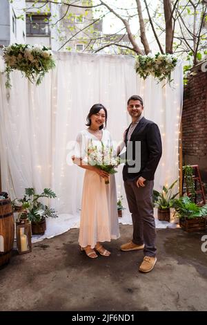 Lächelnde asiatische Braut mit Blumenstrauß in der Hand berühren Bräutigam im Smoking und Blick auf die Kamera, während in der Nähe der Hochzeit Bogen geschmückt stehen Stockfoto