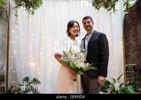 Lächelnde asiatische Braut mit Blumenstrauß in der Hand umarmt Bräutigam in Smoking und Blick auf die Kamera, während in der Nähe der Hochzeit Bogen geschmückt stehen Stockfoto