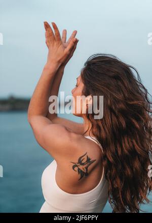 Seitenansicht einer jungen frauenfrauenförmigen Frau mit langen welligen Haaren in Sportswear, die beim Yoga am Meer Mountain mit den Armen nach oben und hinten biegt Stockfoto