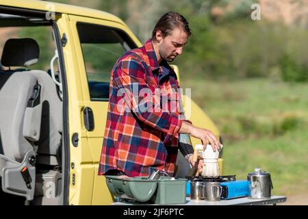 Männliche Reisende in legerer Kleidung Montage Moka Topf auf Gasherd während der Vorbereitung Kaffee zum Frühstück außerhalb gelben Van während der Fahrt in countrysi Stockfoto