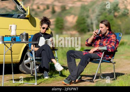 Ganzkörperfrau aus Hispanic in legerer Kleidung und Sonnenbrille beim Lesen eines Buches, während sie auf einem Stuhl neben dem Mann im karierten Hemd sitzt und Kaffee auf dem Campingplatz trinkt Stockfoto