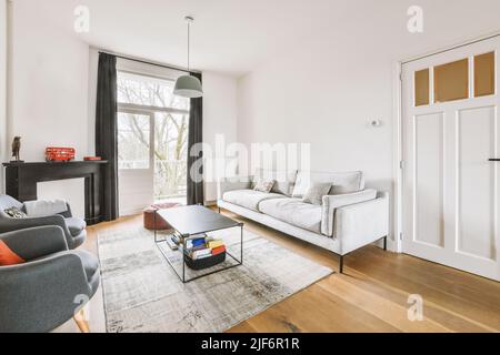 Bequemes Sofa und Sessel auf weichem Teppich in der Nähe der Fenster In gemütlichem hellen Wohnzimmer in moderner Wohnung Stockfoto