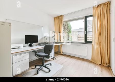 Stuhl am Tisch mit modernem Computer und Drucker an der Wand in hellen Raum mit Vorhängen am Fenster in der Wohnung platziert Stockfoto