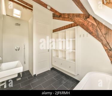 Weiße Badewanne in der Nähe von Waschbecken an der Wand mit Spiegel mit Holzbalkendecke in modernen hellen stilvollen Bad in der Wohnung platziert Stockfoto