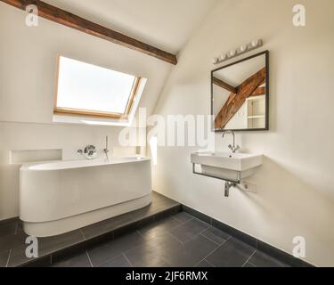 Weiße Badewanne in der Nähe von Waschbecken an der Wand mit Spiegel mit Holzbalkendecke in modernen hellen stilvollen Bad in der Wohnung platziert Stockfoto